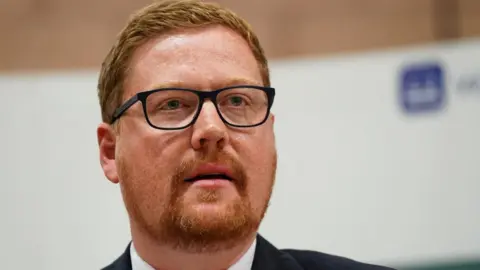 Jonathan Brash, pictured with red hair and beard, looking off to the right. He's in the foreground and a plain, blurred background is behind him. 