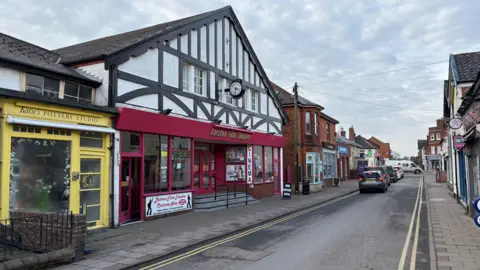 Johnnie Wright/BBC Leiston High Street. There is a yellow pottery studio, next to the cinema which has a burgundy coloured front and glass windows with steps leading up to the entrance. 