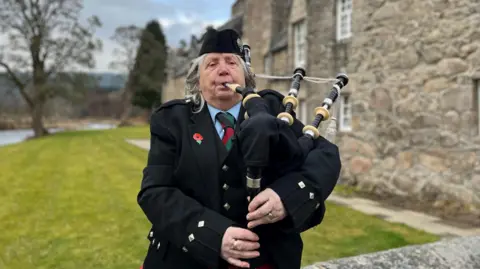 A woman wearing formal Highland dress playing the bagpipes. She has grey hair and is wearing a  black jacket. There are an old building and trees in the background.