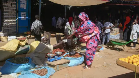 AFP A woman wearing a patterned pink and blue dress buys from a market trader, who is sat on the floor at Abu Shouk's market, surrounded by produce.