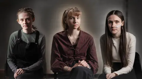 Three teenage girls sitting in front of a grey background.