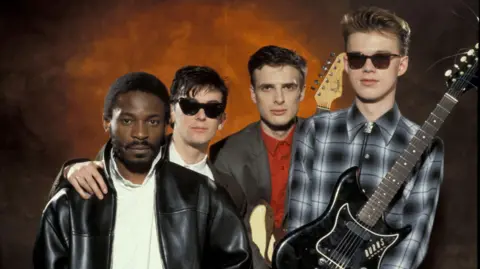 The band Orange Juice - Zeke Manyika (wearing a black leather jacket and white shirt), James Kirk (wearing shades) David McClymont (grey suit and red jacket) and  Edwyn Collins (chequed shirt, holding a black guitar) look into the camera