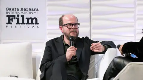 Getty Images Nick Emerson speaking at the Santa Barbara International Film Festival. He is sitting on a white sofa, with white boards behind him. He is wearing a dark suit with open neck shirt. 