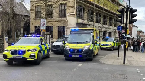 A police car, a police van, an ambulance and an ambulance van parked on Bank Plain in Norwich. A large group of people are standing to the right side of the vehicles. a building in the background is covered in scaffolding.