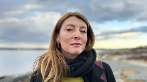 BBC A woman with long red hair wearing a black scarf and coat looks at the camera. Behind her is the sea and a rock