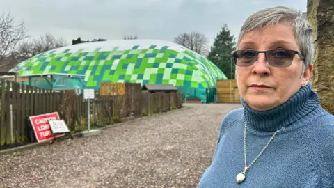 Jacquie Christie is standing near the green giant inflatable. She has grey short hair and glasses. She is wearing a blue jumper with a silver pendant necklace over the top.