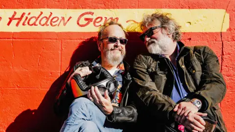 South Shore Productions Dave Myers and Si King sit in front of a painted orange wall. They are both wearing biker jackets and have sunglasses on. Dave is smiling contentedly and Si is looking at Dave with fondness. Behind them, written on the wall, is a sign that reads "hidden gem".