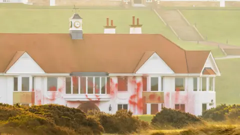 Red paint covering the front of the Trump Turnberry resort. The building is white with an orange roof. It has red paint covering part of the frontage and windows.