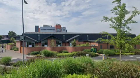 Google A single-story redbrick building with a black roof surrounded by bushes and trees and a road running outside it. In the background another building and above a blue sky with some clouds.