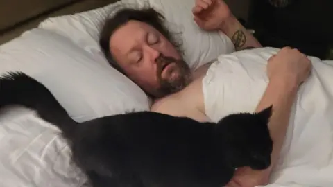 A man laying in bed with his eyes closed with the black cat next to him