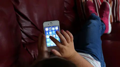 A child lies on a red sofa and is scrolling along a mobile phone screen with their finger 