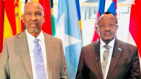 Somalia's Family Ministry / X Gen Jama (R) with former MP Abdullahi Godah Barre (L) pictured in front of the Somali flag - and other flags - at the UN meeting. Both are wearing suits and ties and look serious.