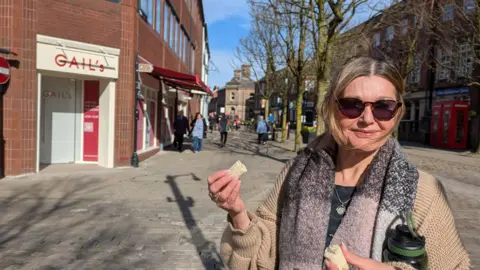 Jane Kent in sunglasses in front of Gail's shopfront eating a banana