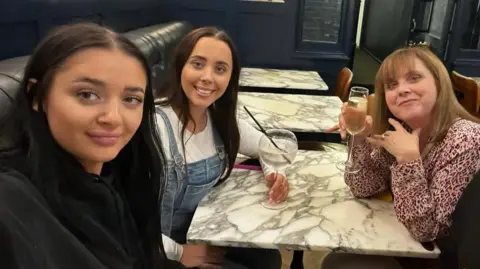 Emma Ledgard An image of Carol, Louise and Hannah Hunt sitting in a restaurant and smiling. From right to left, Louise has long black hair, is wearing black and on the right. Hannah has long dark hair and is wearing a white top under blue dungarees. Carol has light brown hair and is wearing a pink top with a small dark print.