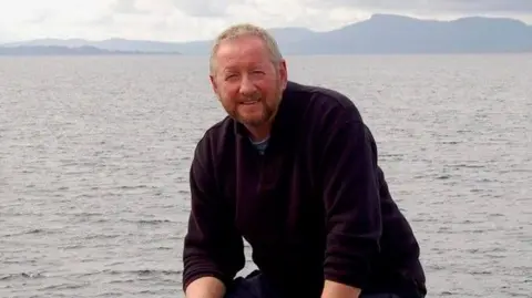 Alan Beattie Herriot Alan Beattie Herriot is sitting on a rock with the sea behind him. He has short hair and a beard.