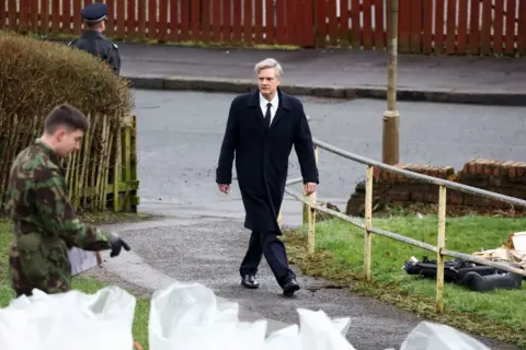 Getty Images Colin Firth on set in character as Dr Jim Swire - he is walking up a pathway looking concerned an is wearing a long dark coat, a white shirt and a black tie. There is an army officer in the foreground and a police officer in the background.