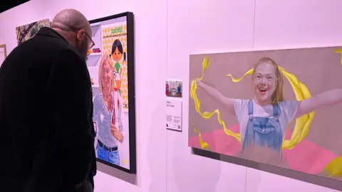 Tom Ingall/BBC A bald man with a beard reads a plaque next to a painting of a young girl smiling with her arms outstretched on a gallery wall. 