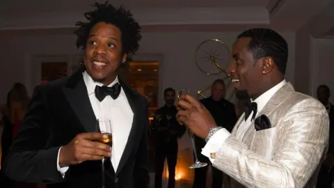 Getty Images File image of Jay-Z and Diddy at an event - Jay Z is dressed in black tie and is smiling and looking off to his right, while Sean Combs, on the right, is looking across at him and smiling. He is wearing black tie, but with an shiny ivory coloured jacket. Both men are holding champagne flutes. 