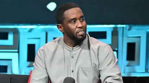 Getty Images Sean "Diddy" Combs is photographed during an interview. He is sitting in a tan button-down shirt and holding a microphone. 