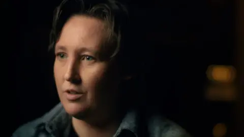 Mhairi Black is dressed in a grey shirt and is against a black background. She is almost in profile facing to the left of the picture.