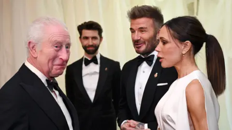 PA Media King Charles speaks with David and Victoria Beckham. Both men wear black tuxedos, while Victoria Beckham wears a sleeveless white gown and has her brown hair tied up in a ponytail.