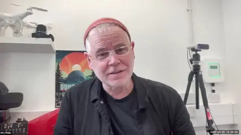 A man with an orange hat and black shirt sits beside drones and a camera on a tripod.