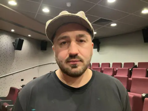 Kostiantyn Bidnenko wearing a hat looks at the camera with red seats behind him