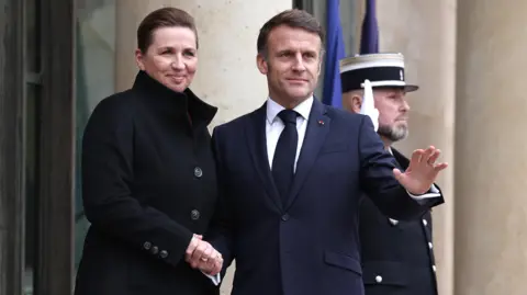 Mette Frederiksen, wearing a dark coat, shakes President Macron's hand. He is wearing a dark blue suit jacket and a tie.