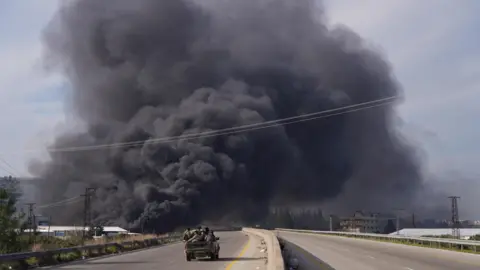 Image from Friday shows thick black smoke rising in the distance as Syrian forces riding in a pickup truck drive towards it on an empty road