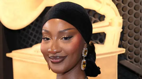 Getty Images A headshot of Tems wearing a black headscarf smiling and looking at the camera with a large image of golden Grammy behind her.