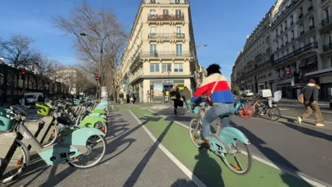 Cyclist in red, white and blue coat rides along green cycle lane in Paris next to a row of rentable bikes