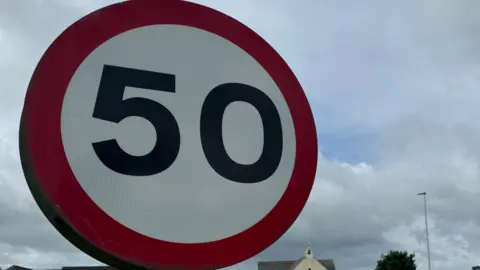 BBC On the left of the image is a circle which has a red boarder around it. The middle part is white, and has a black number 50 written on it. It is a road sign that is on the side of a road. 