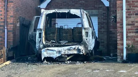 A burned out jeep sits in the drive way of a house. it is seen from behind looking into the boot and is completely destroyed by fire. A garage stands behind it while the driveway is covered in detritus 