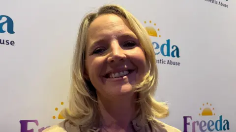 Kate Wright is standing in front of a banner with the Freeda logo on it, which includes a sunrise. She has shoulder-length blonde hair and is smiling at the camera.