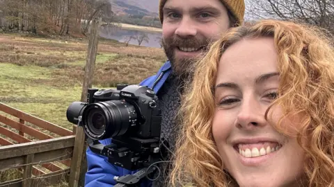 Ciara Hardisty, who has with strawberry blonde wavy hair, is in the fore, with Adam Morgan, who is wearing a hat, has a beard and is holding a black camera, to the left behind her. There are fields, trees and a lake behind them in the distance.