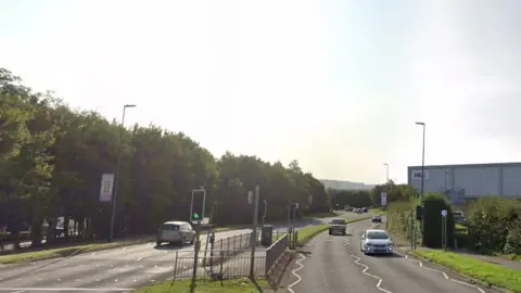 Marsham Way in Longwell Green. Cars are going in both directions on the road, there is a M&S on the right side and trees on the left.