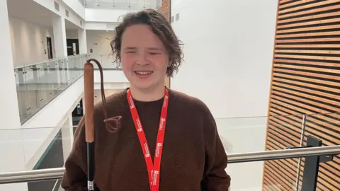 Annabel Amos/BBC A girl with long brown hair wearing a brown jumper and a red University of Northampton lanyard stands in a stairwell holding a cane.