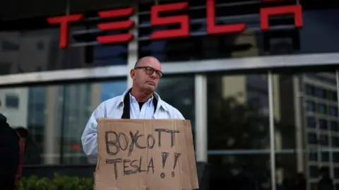 Reuters A man holds a sign - made from what looks like a scrap piece of cardboard - which reads "Boycott Tesla"