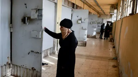 Reuters A woman opens a prison door in a corridor of Saydnaya prison.  She is wearing a dark coat and hat and her face is covered with a scarf.  In the background, along the corridor, is another group of people.