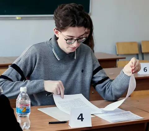 Lik family  Kevin sitting at a desk looking at a test paper, wearing black glasses and a grey jumper - he has thick dark hair