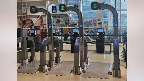 Three giant scales in front of Tesco Scan as you Shop checkouts. They have a large metal plate on the ground and barriers either side with numbers one to three above. Reddit users have posted that they look like airport security scanners.