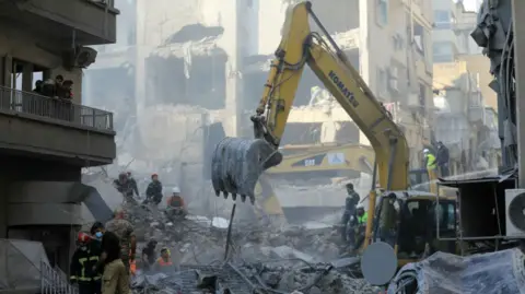 The scene of destruction following a massive Israeli strike on central Beirut. An excavator is digging through the rubble as dozens of rescue workers move around. It's daylight and dust hangs in the air.