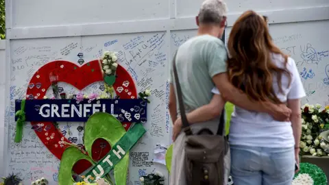 Getty Images Two people hug in front of the Grenfell remembrance wall, with a heart in the background that says 'Grenfell' attached to the wall and messages people have written on the wall, in London 