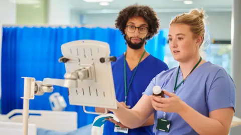 Two hospital workers dressed in blue scrubs looking at what appears to be a clipboard. The female worker, who has pulled-backed blonde hair, is holding a bottle of medication. The male worker who has curly dark hair and a beard looks on.