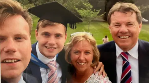 Family handout Mr O'Sullivan, pictured on his graduation day with his family.  His brother is on the left of the frame. He has fair short hair and is smiling. Jack is next to him. He also has short fair hair and is wearing a graduation cap and gown over a white shirt and pink and blue striped tie. Catherine is next to him. She has short blonde hair and has glasses on her head. She is looking at the camera and smiling. Alan O'Sullivan is on the right and is wearing a suit.