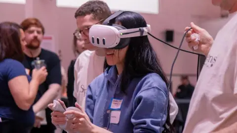 A woman wears a virtual reality headset while holding a controller in each hand. Two people are standing by her and watching, while three people are blurred out as they talk to each other in the background.