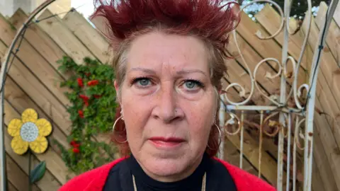 Jenny Carruthers looking into the camera with a concerned expression. She is wearing a black turtle neck, a red cardigan and gold chain. She has burgundy hair styled upwards in spikey points and is wearing hoop earrings and a nose stud. She is standing in her garden with a wooden fence, greenery trellis, and a flower mosaic decoration behind her.