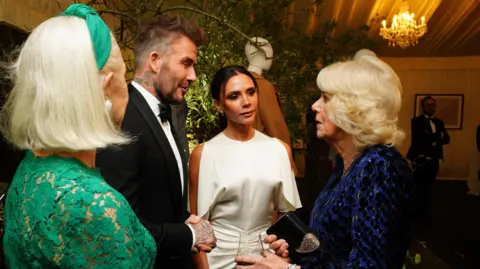 PA Media Queen Camilla stands in a huddle with the Beckhams and Dame Helen Mirren. The Queen wears a black, long-sleeved dress with a blue and green pattern. Victoria Beckham wears a sleeveless white dress with ruching at the hips. David Beckham wears a black tuxedo. Dame Helen Mirren wears a long-sleeved green dress with a lace top.