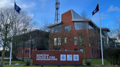 Three-storey brick building with glass around the top floor. There are several antennae on a pole on top of the building, and another glass and brick building to the left. A sign showing "Norfolk Police and Fire Headquarters" is visible on grass in the foreground.