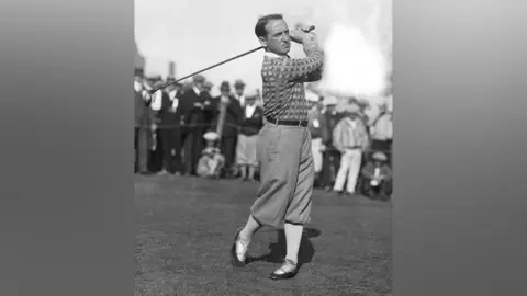 USGA Museum Old black and white image of golfer, Bobby Cruickshank, swinging golf club, with trousers tucked into socks, with crowd of smartly-dressed spectators in the background.